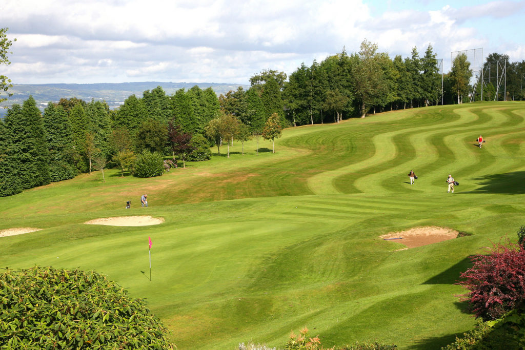 A lush green golf course fairway with golfers in the distance, overlooking a sparkling body of water (Belfast Lough) from Holywood Golf Club, Holywood, Northern Ireland.